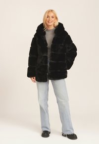 Black faux fur coat with a hood, featuring a boxy design and wide sleeves. Worn over a gray ribbed top and paired with light wash jeans.
