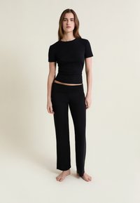 Jeune femme debout pieds nus, portant un haut noir ajusté à manches courtes et un pantalon large noir, sur un fond clair uni.