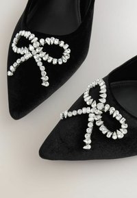 Fekete bársony, hegyes orrú balerina cipők, nagy, átlátszó rhinestone-ből készült masni díszítéssel, különböző formákban és méretekben, dekoratív akcentusokként.