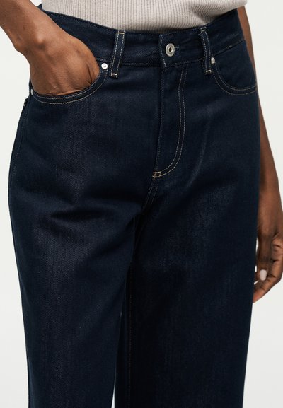 Tamsiai mėlynos spalvos džinsai iš džinso audinio su matomomis siūlėmis, sagomis ir užtrauktuku, dėvimi asmens, kurio viena ranka kišenėje ir dėvi smėlio spalvos viršutinę dalį.