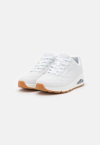 White sneakers with a smooth synthetic upper, perforated details, and light brown rubber outsole. Features lace-up closure and visible air cushioning.