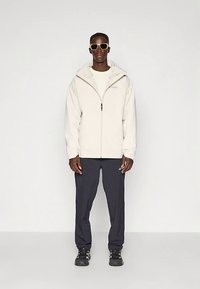 Svijetlo bež jakna s patentnim zatvaračem i kapuljačom, uparena s tamnim hlačama i crnim cipelama. Ima minimalistički dizajn i vidljiv logo na prsima.