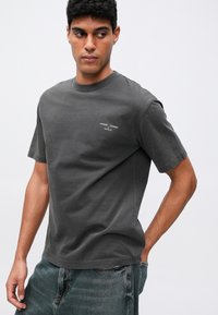 Camiseta de algodón gris con mangas cortas, cuello redondo y pequeño texto gráfico blanco en el pecho. Combinada con pantalones cortos de mezclilla oscuros.