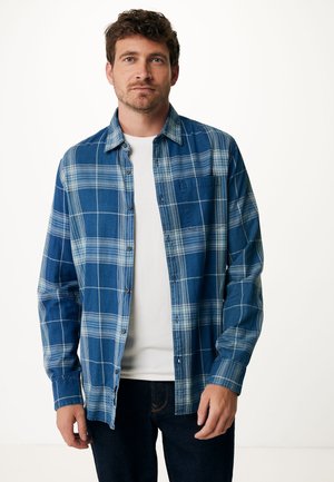 Camisa de franela a cuadros azules con cuadrados en azul claro y azul oscuro, con botones en la parte delantera, un bolsillo en el pecho, usada sobre una camiseta blanca.