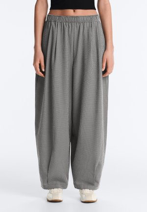 Žena u širokim, visokim crno-bijelim kariranim hlačama širokih nogavica i bijelim tenisicama, u kratkoj crnoj majici.