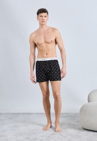 Άνδρας χωρίς πουκάμισο, όρθιος ξυπόλυτος σε χαλί, φοράει μαύρα αντρικά εσώρουχα Calvin Klein με λευκή ζώνη μέσης με λογότυπο και μικρά λευκά σχέδια.