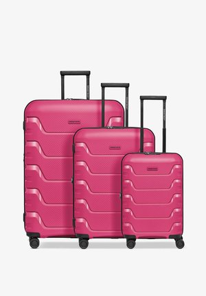 Trois valises rigides roses de tailles différentes avec des poignées télescopiques et quatre roues pivotantes chacune, disposées côte à côte.