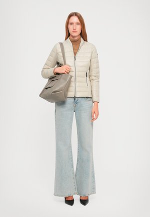 Donna che indossa un giubbotto imbottito beige, maglione a collo alto marrone, jeans a zampa d'elefante azzurri chiari, scarpe nere a punta, mentre tiene una borsa grigia su sfondo bianco.