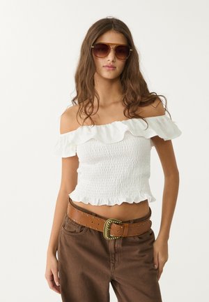 Top bianco off-the-shoulder con scollatura a volants e texture arricciata, abbinato a pantaloni marroni a vita alta e una cintura beige con borchie.