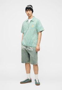 Νεαρός άνδρας που φοράει μαύρο και λευκό σκουφάκι, πράσινο ριγέ κοντομάνικο πουκάμισο, τζιν σορτς με λευκό γραφικό, λευκές κάλτσες και πράσινα αθλητικά παπούτσια.