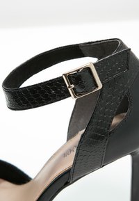 Soulier noir à talons hauts avec une sangle à motif serpent texturé, fermeture à boucle et accents en cuir lisse. Présente un côté ouvert pour une meilleure respirabilité.