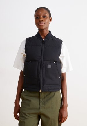 Black padded vest with a high collar, front zipper, two large pockets, and small logo tag. Worn over a white shirt and olive pants.