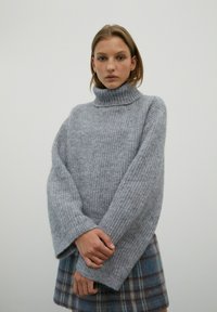 Chunky grauer Strickrollkragenpullover mit weiten Ärmeln und gerippter Textur, kombiniert mit einem karierten Rock in Blau- und Brauntönen.