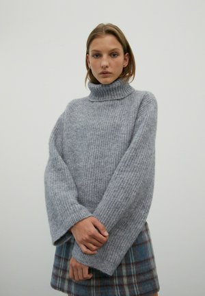 Dikke grijze gebreide coltrui met wijde mouwen en ribbelstructuur, gedragen met een geruite rok in blauwe en bruine tinten.