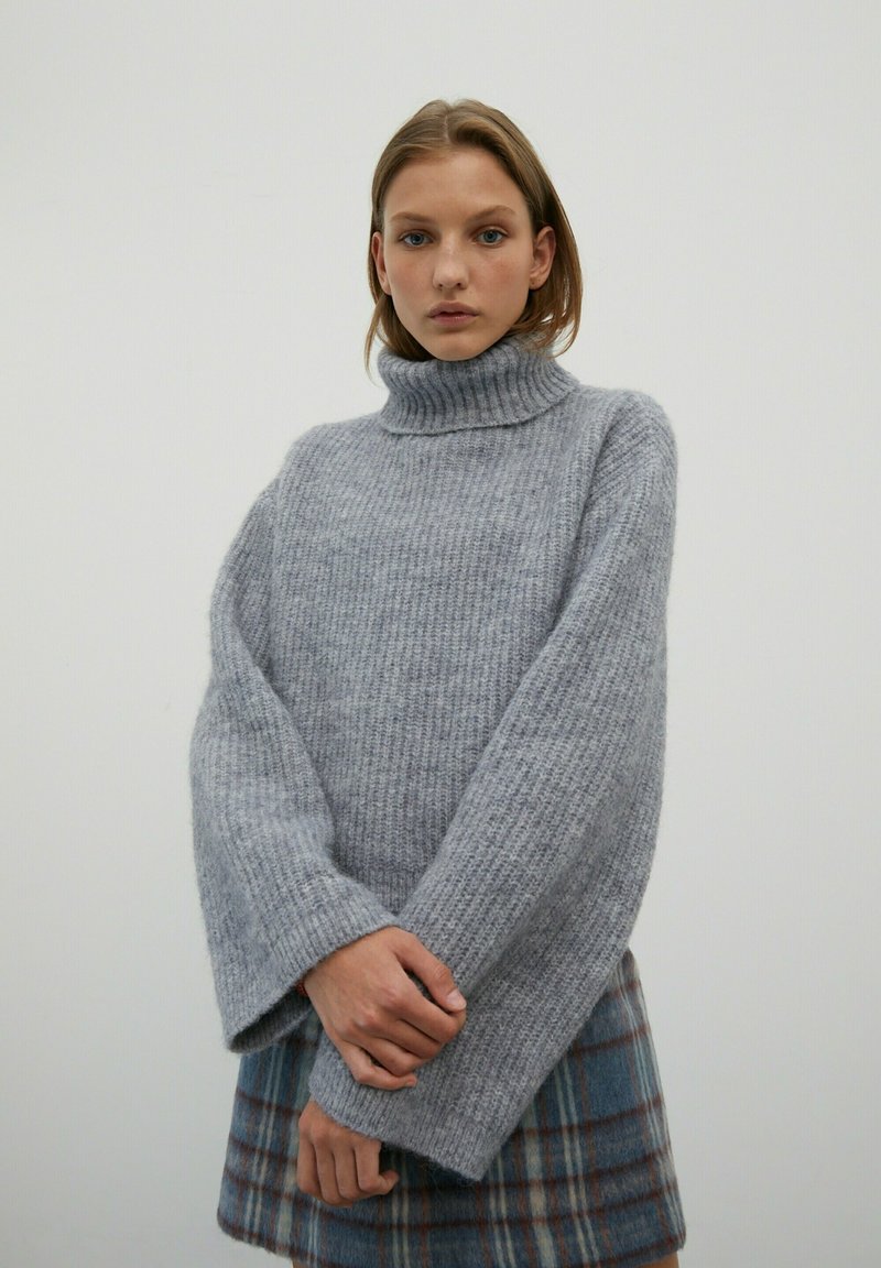 Pull en maille épaisse gris avec col roulé, à manches larges et texture côtelée, associé à une jupe à carreaux dans des tons de bleu et de marron.