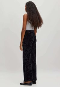 Pantalon large en velours noir avec une texture lisse et une taille haute, associé à un haut gris clair. Modèle vu de dos.