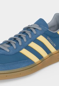 Μπλε παπούτσι Adidas Spezial με κίτρινες ρίγες, γκρι κορδόνια και λαστιχένια σόλα, που δείχνει λεπτομέρειες από το πλάι και από πάνω.