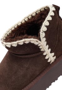 Bruine suède enkelboot met een zachte bontvoering, voorzien van een decoratieve crème kleurige stiksel langs de opening en een gestructureerde rubberen zool.
