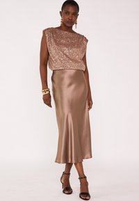 Top bronzo decorato con paillettes e scollatura a barca abbinato a una gonna satinata bronzo di lunghezza media. Sandali neri con cinturini completano l'outfit.