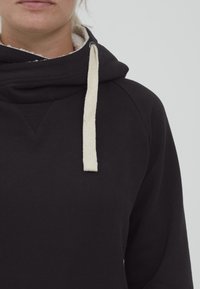 Sudadera negra con interior de forro polar, que presenta un acento de punto triangular en el cuello y un cordón de color crema.