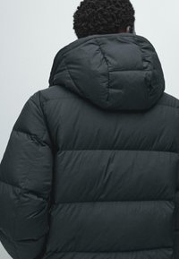 Persona con capelli ricci corti che indossa un giubbotto imbottito nero con un grande cappuccio, vista di spalle contro uno sfondo neutro.