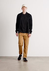 Ung mand går iført beige bukser, sort jakke, sort skjorte, sorte sneakers og hvid baseballcap mod en ensfarvet baggrund.