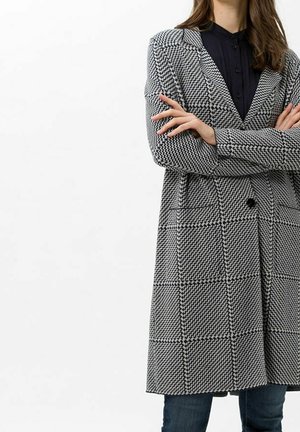 Femme portant un manteau à carreaux noir et blanc par-dessus une chemise noire, les bras croisés, debout devant un fond blanc uni.