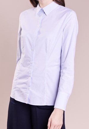 Woman wearing a light blue long-sleeve button-up shirt with a pointed collar and black pants, standing against a plain background.