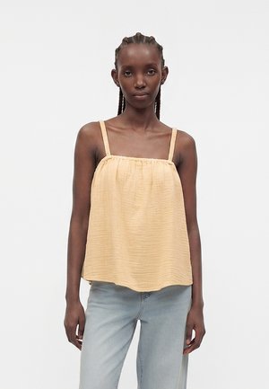 Γυναίκα με πλεκτά μαλλιά, που φοράει ένα κίτρινο αμάνικο κορσέ με υφή και ανοιχτό μπλε τζιν, στέκεται μπροστά σε λευκό απλό φόντο.