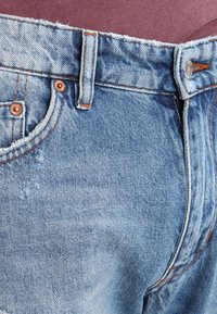 Zbliżenie na talię jasnoniebieskich dżinsów, z miedzianymi nitami, pomarańczowymi szwami i srebrnym guzikiem, na różowym tle.