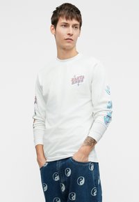 Jauns vīrietis, tērpies baltā garām piedurknēm kreklā ar krāsainiem attēliem un zilos džinsos, kas aprakstīti ar jingi un jangs simboliem, rokas kabatās.