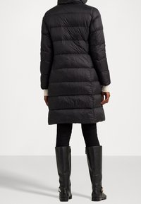 Svart puffjacka med lång silhuett, quiltat mönster, hög krage och ribbade ärmslut. Kombinerad med figursydda svarta leggings och knähöga stövlar.