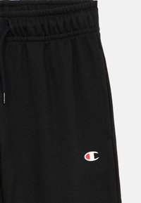 Μαύρο παντελόνι φόρμας από μαλακό ύφασμα, με ελαστική μέση και ένα μικρό κόκκινο και λευκό λογότυπο στην αριστερή πλευρά.