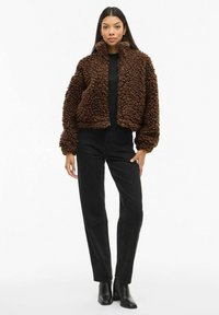 Smeđa jakna od umjetnog krzna s visokim ovratnikom, patentnim zatvaračem sprijeda i bujnim rukavima, uparena s crnim hlačama visokog struka i čizmama do gležnja.