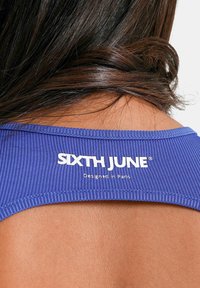 Lilla ribbet stof med et logo mærke, der læser "SIXTH JUNE" og "Designet i Paris", sat mod glat hud.