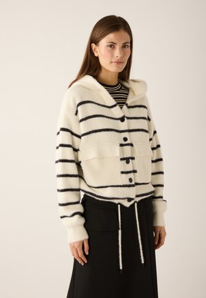 Weißer, flauschiger Kapuzenpullover mit schwarzen Streifen, vorne mit Knöpfen und zwei Taschen. Getragen über einem schwarzen Rock mit Kordelzügen.