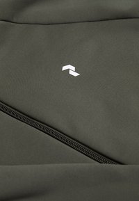 Green zippered fabric with a smooth texture features a small white logo resembling an abstract design. The overall look is minimalist.