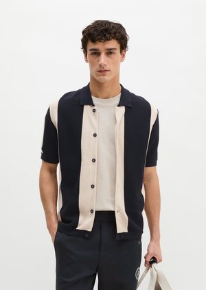 Junger Mann trägt ein schwarz-beiges, vertikal gestreiftes Hemd mit Knöpfen über einem weißen T-Shirt, schwarze Hose, hält eine beige Tragetasche.