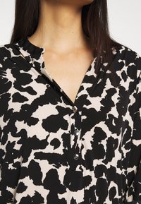 Svart och gräddfärgad blus med leopardmönster, med krage och två silverknappar vid halsringningen. Slät tyger.