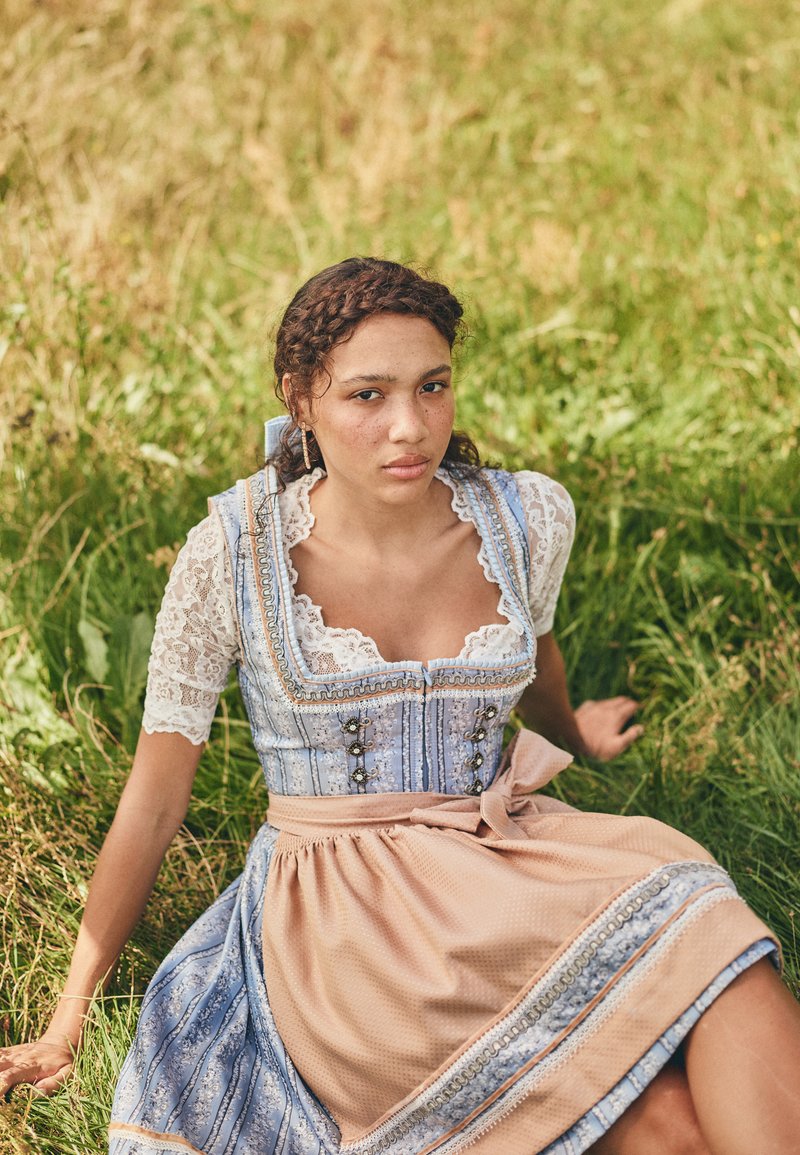 Blau-weiß gemusterter Dirndl mit Spitzenärmeln, tailliertem Oberteil und einem beigen Rock, der mit einem Satinband und dekorativen Knöpfen akzentuiert ist.
