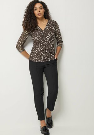 Kvinde med krøllet hår iført en brun mønstret V-hals top, sorte smalle bukser og sorte loafers foran en hvid væg.