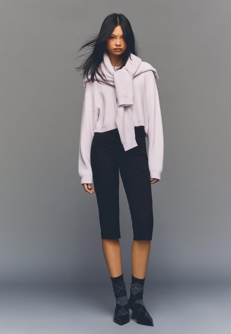 Skrčen ogrnjen pulover iz sivke, slojen z ujemajočo se ruto, združen s črnimi hlačami do kolen in vzorčastimi črnimi gležnjnimi nogavicami ter petami.