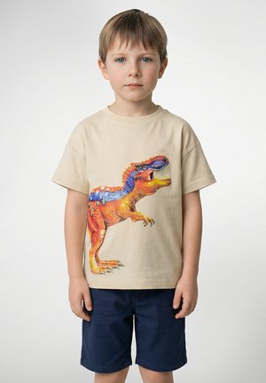 T-shirt beige in cotone con grafica colorata di un dinosauro. Maniche corte, scollo rotondo, vestibilità rilassata. Abbinata a pantaloncini blu navy.