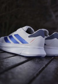 Chaussures de sport blanches avec des rayures bleues, dotées d'un matériau supérieur texturé, d'un col rembourré et d'une épaisse semelle blanche avec un accent bleu.