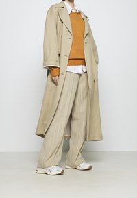 Trench coat beige sopra a un maglione arancione, camicia bianca e pantaloni a righe beige. Completato con sneakers bianche e beige.