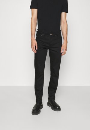 Mężczyzna ubrany w czarne obcisłe dżinsy, czarną koszulkę i czarne skórzane buty, stojący na tle jednolitego jasnego tła.