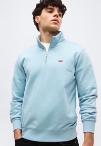 Νεαρός άνδρας που φοράει ανοιχτό μπλε φλις με φερμουάρ μέχρι τη μέση και μικρό κόκκινο λογότυπο Levi's, με τα χέρια στις τσέπες, κοιτώντας ελαφρώς προς τα πάνω και πλάγια.