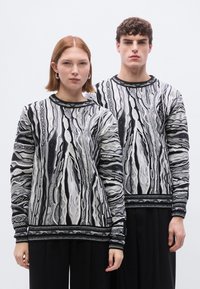 Pulls en maille à manches longues noir et blanc avec motifs ; présentent des rayures texturées et des dessins complexes ; portés avec un pantalon noir.