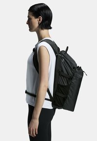 Black backpack with adjustable straps, textured panels, drawstring closure, and side pockets, worn on a person in a white shirt.