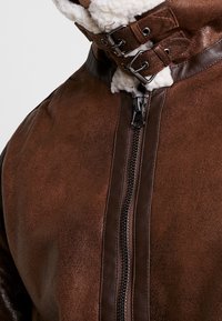 Bruine leren jas met een gestructureerde afwerking, voorzien van een ritssluiting aan de voorkant en een dubbele gesp aan de kraag. Gevoerd met crèmekleurig schapenvacht.